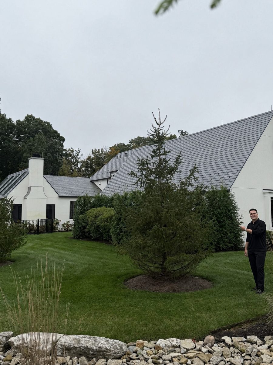 Damon Coon of Pink Tree Care gesturing toward a young spruce in a landscaped yard beside a white house